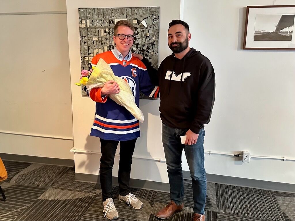 Chris Buyze, wearing an Edmonton Oilers jersey, holds a bouquet of pink and yellow flowers and stands beside a smiling Anand Pye
