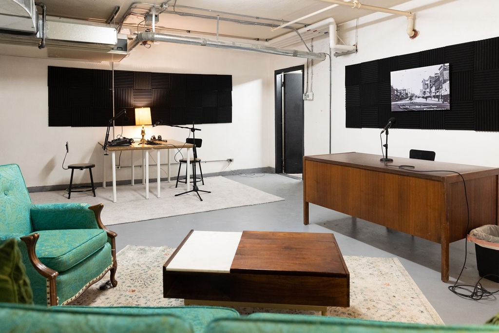 The inside of Homestead Coworking's podcast vault, featuring green furniture, a table with microphones and chairs, a desk with another microphone, a coffee table, and black foam panelling on the walls