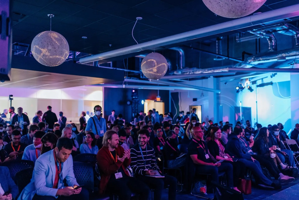 A large crowd sits in rows under blue and purple lights while awaiting a presentation during Amii's first AI week.