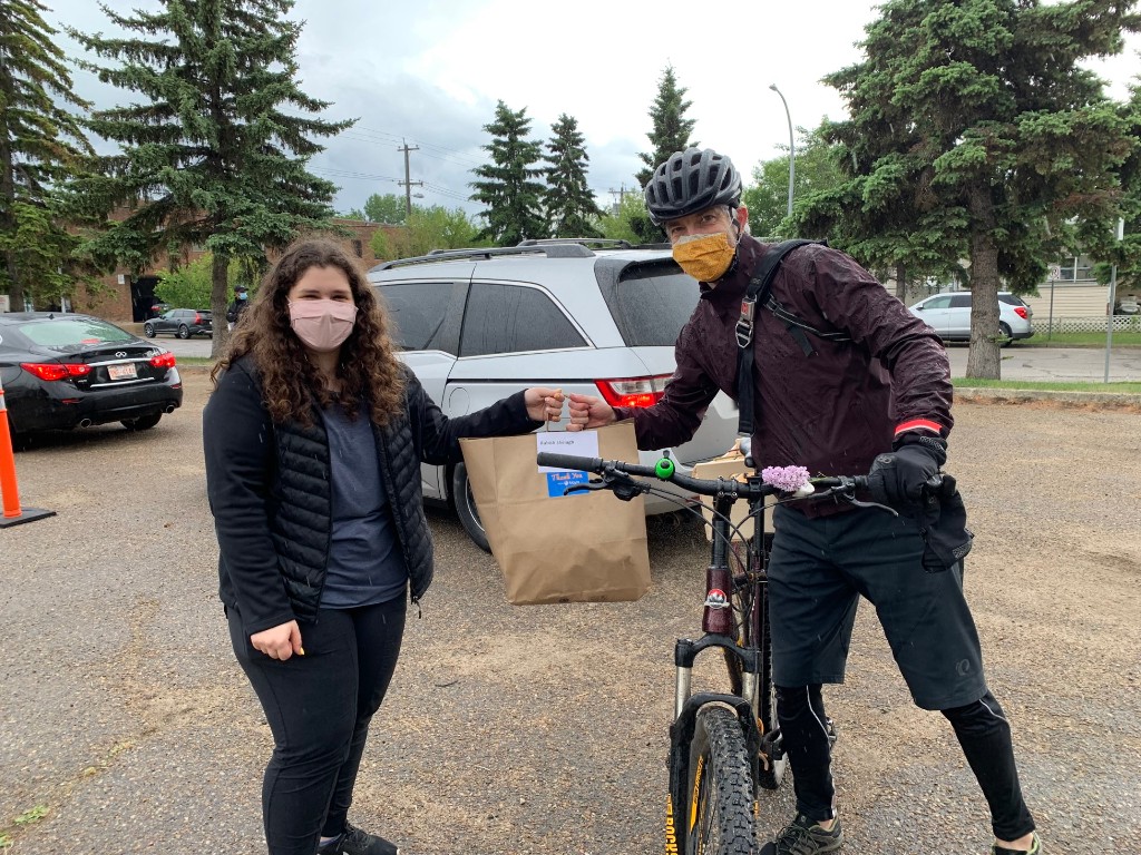A masked man on a bike accepts a paper bag of bagels from a young woman in a parking lot