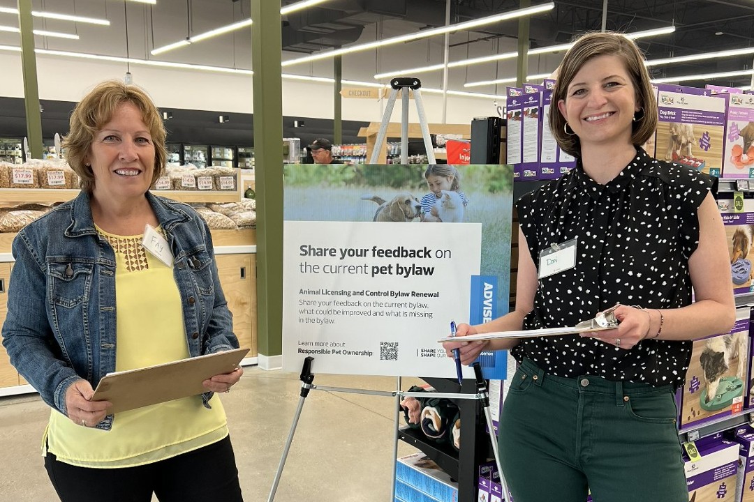 Two women with clipboards stand in a pet store in front of a sign that reads "Share your feedback on the current pet bylaw"