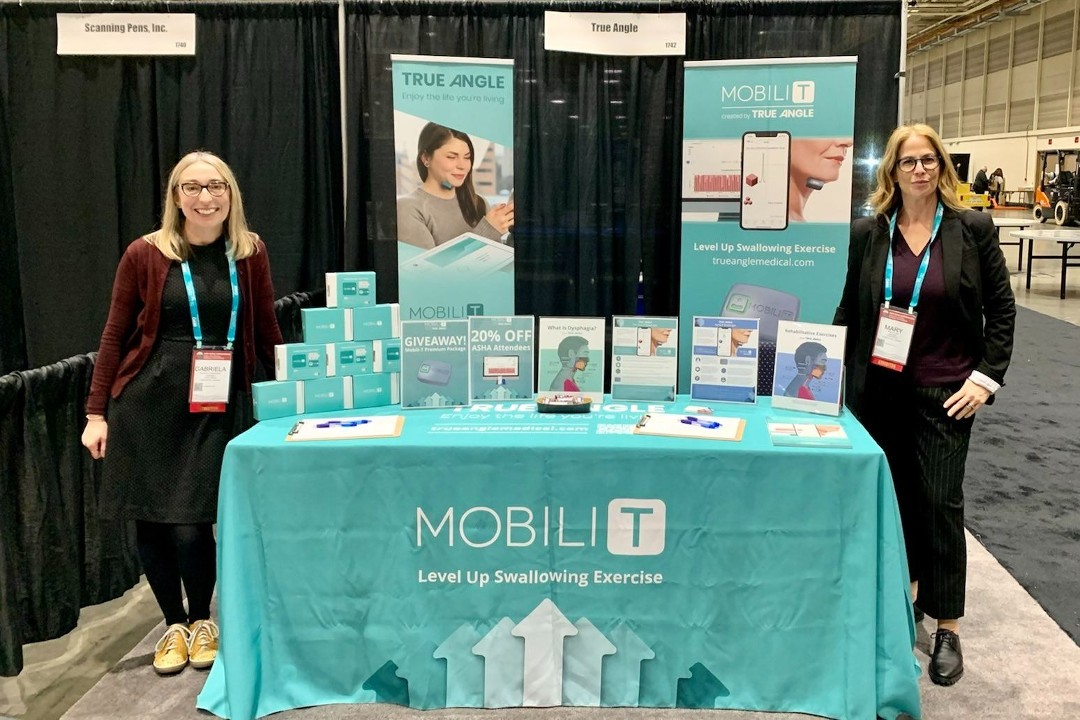 Two women wearing lanyards stand beside True Angle's booth at a trade show, displaying the Mobili-T device