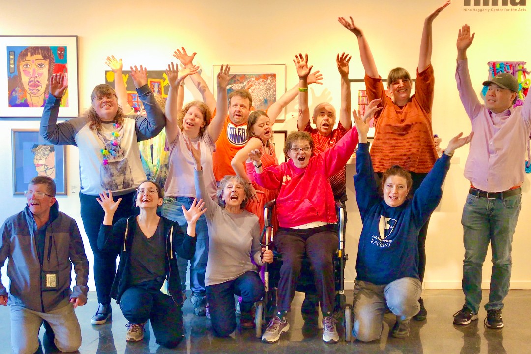 Twelve people express joy with big smiles and arms stretched high. Behind them is a wall of paintings at the Nina Haggerty Centre for the Arts.