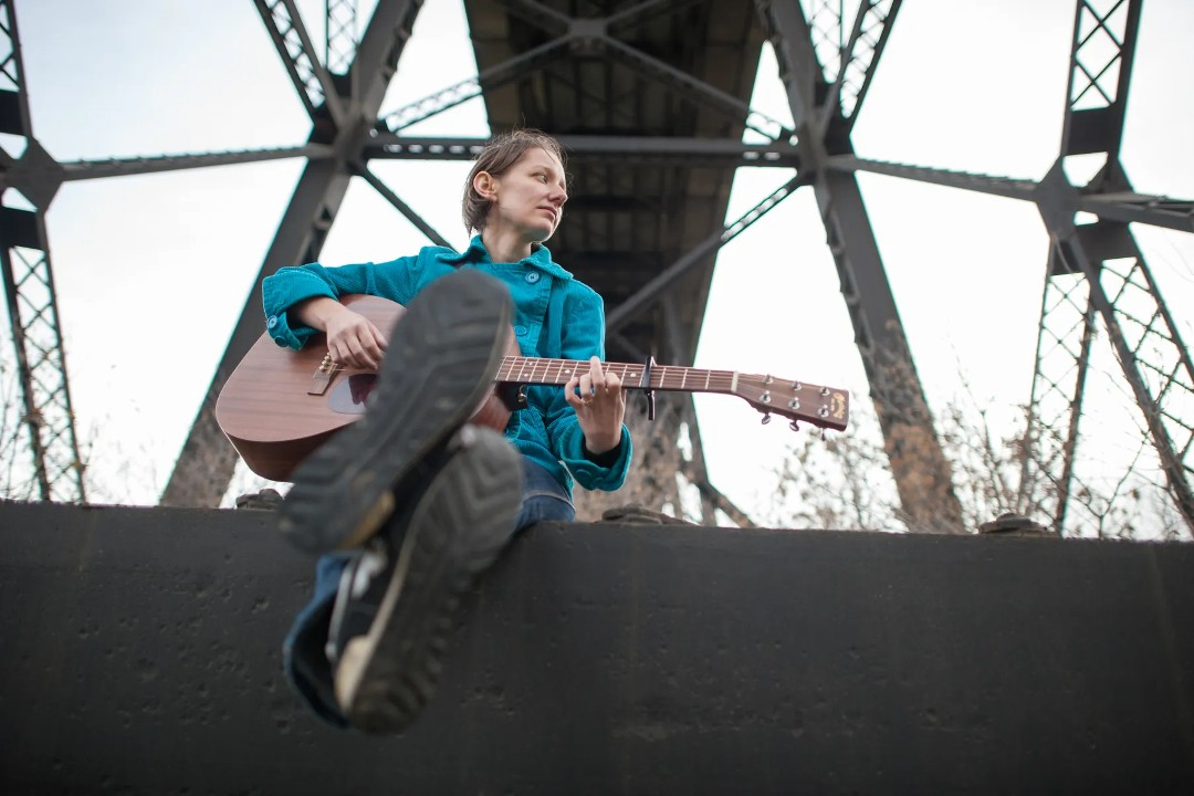 A woman, Dana Wylie, sits underneath a bridge, holding a guitar