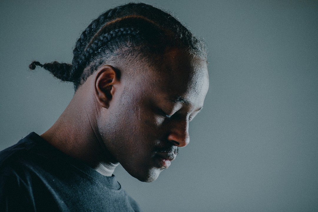 Rapper Mouraine poses in profile with his eyes closed. He is wearing a simple black T-shirt and his hair is braided.