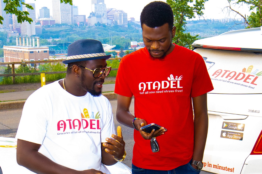Ayomikun Kayode, left, and Temi Kayode, right, look at a cellphone while wearing shirts with Atadel branding, with Edmonton's river valley in the background
