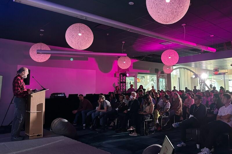 A bearded man speaks at a podium to a full audience in a pink-lit room