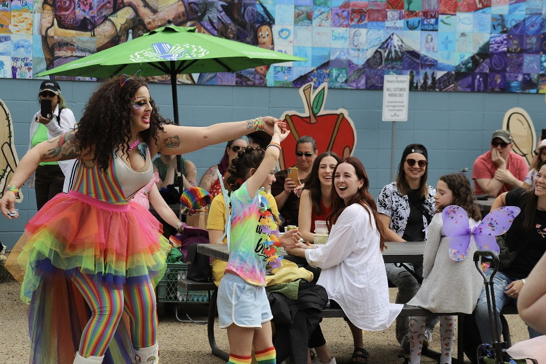 A dancer in a rainbow-hued leotard and a rainbow tutu twirls with a girl wearing Pride socks and a lei at an outdoor event