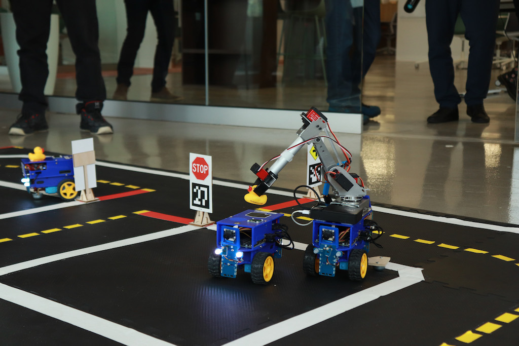 A grey robotic arm lifts a yellow rubber duck off of a blue, boxy, robot with wheels. They are on a mat that looks like a city street.