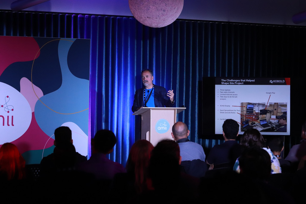 David Cooper of Kobold is speaking at a podium in front of blue curtains, and wears a dark blue suit. There is a powerpoint presentation to his right.