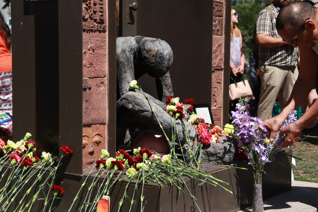 A person lays a flower among many others on a sculpture of a faceless figure, hunched over in a doorway.
