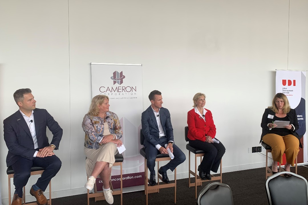 Four panellists and one moderator are seated atop barstools with two chairs in the foreground and two banners in the background of the photo. Each person depicted wears business casual attire.