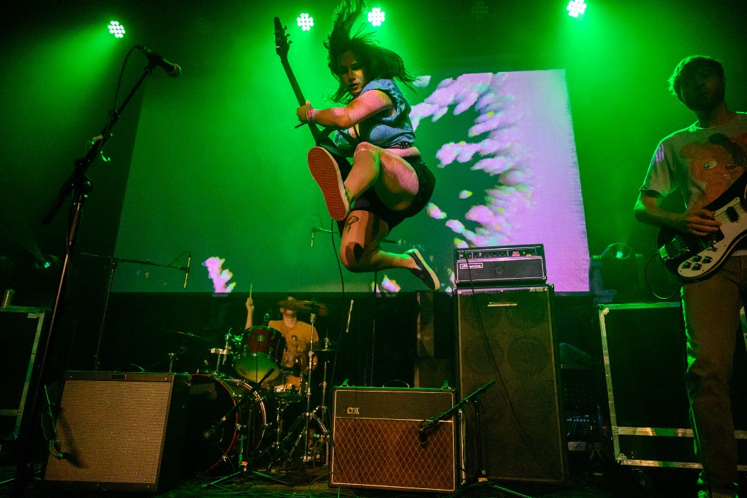 A drummer, guitarist, and bassist perform on stage under green lights. In the centre, the guitarist is mid-leap with her legs kicked out in opposite directions.