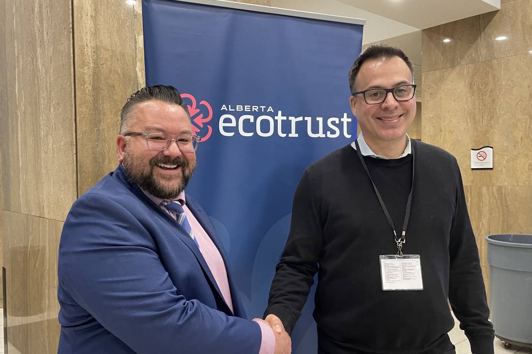 Two men, one in a blue suit and the other in a black sweater, smile and shake hands in front of a banner that reads "Alberta Ecotrust"