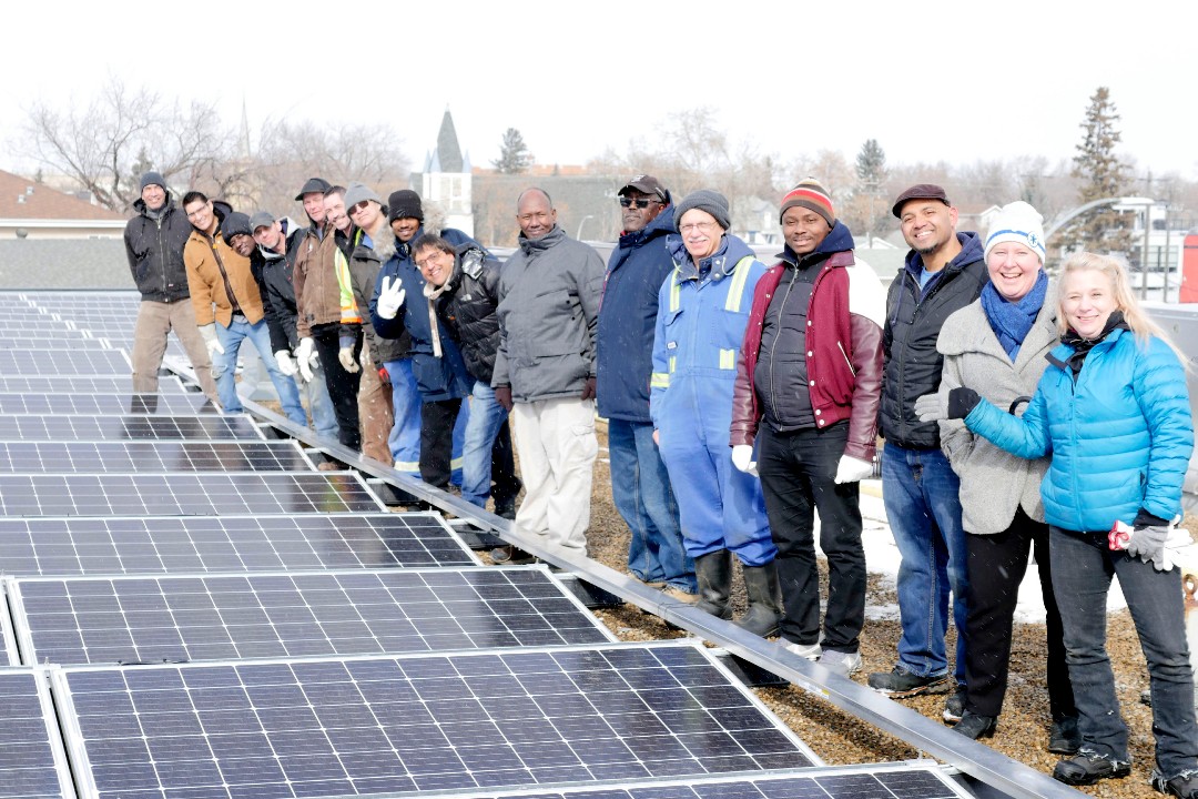 A large group of people stand to the right of a solar panel array on a rooftop. The sky behind them is cloudy and grey.