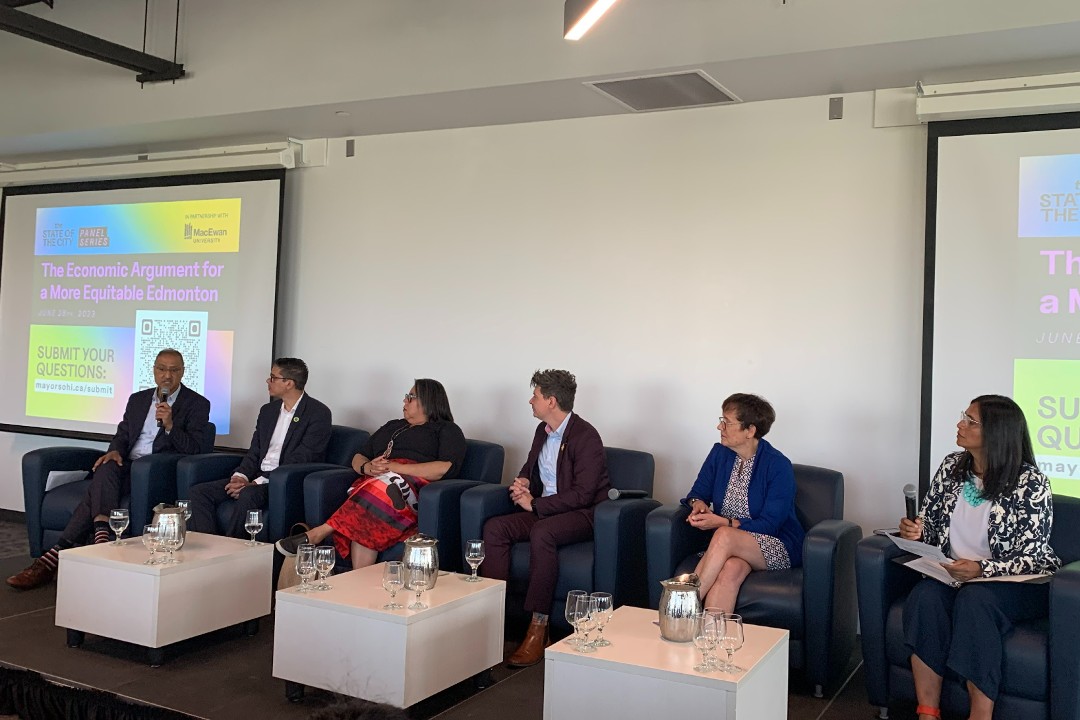 Six people sit on upholstered chairs on a stage, flanked by screens that read "The Economic Argument for a More Equitable Edmonton"