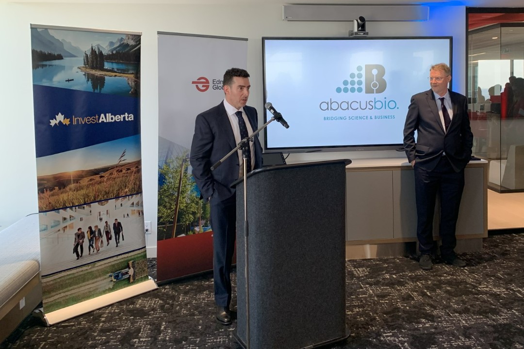 John Crowley speaks at a podium while Peter Amer listens. Behind them are banners for Invest Alberta and Edmonton Global, with the AbacusBio logo projected on a screen.