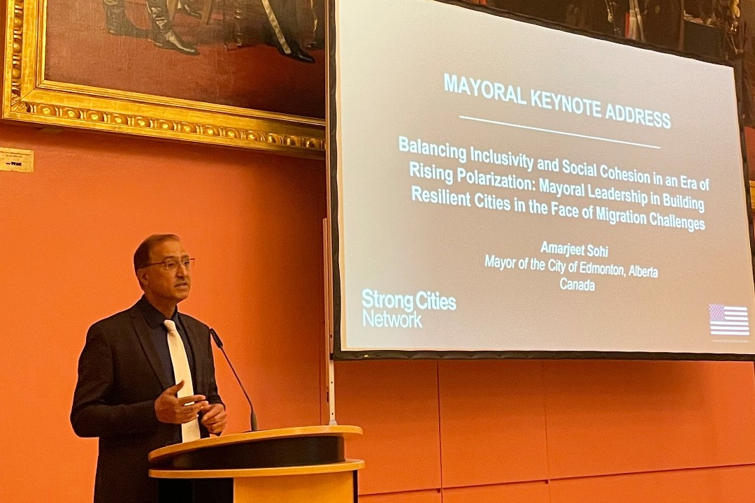 Mayor Amarjeet Sohi speaks at a podium beside a slide that reads "Mayoral Keynote Address: Balancing Inclusivity and Social Cohesion in an Era of Rising Polarization: Mayoral Leadership in Building Resilient Cities in the Face of Migration Challenges"