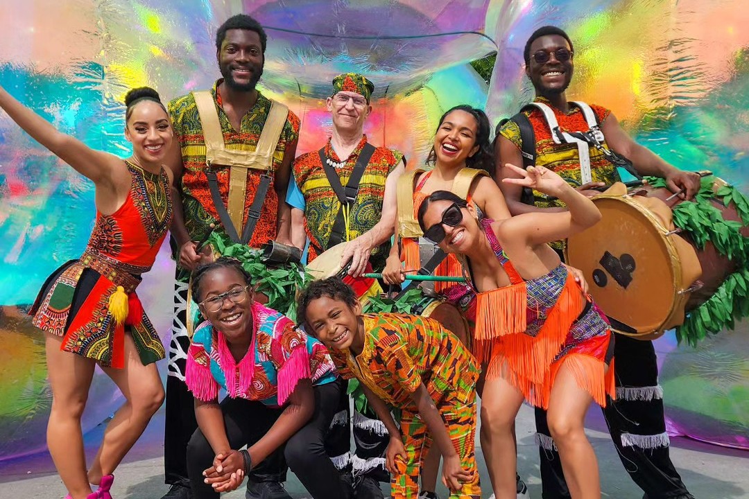 Eight people dressed in bright colours smile and laugh; some are holding drums.