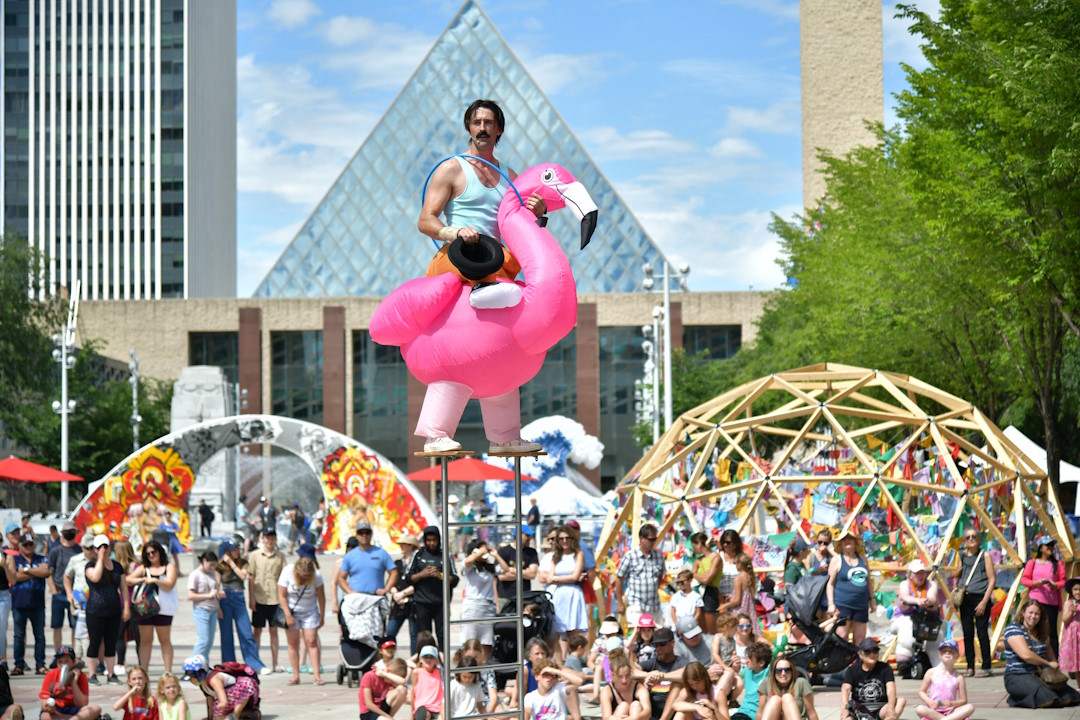 A performer on stilts with an inflatable flamingo surrounded by a crowd with City Hall in the background