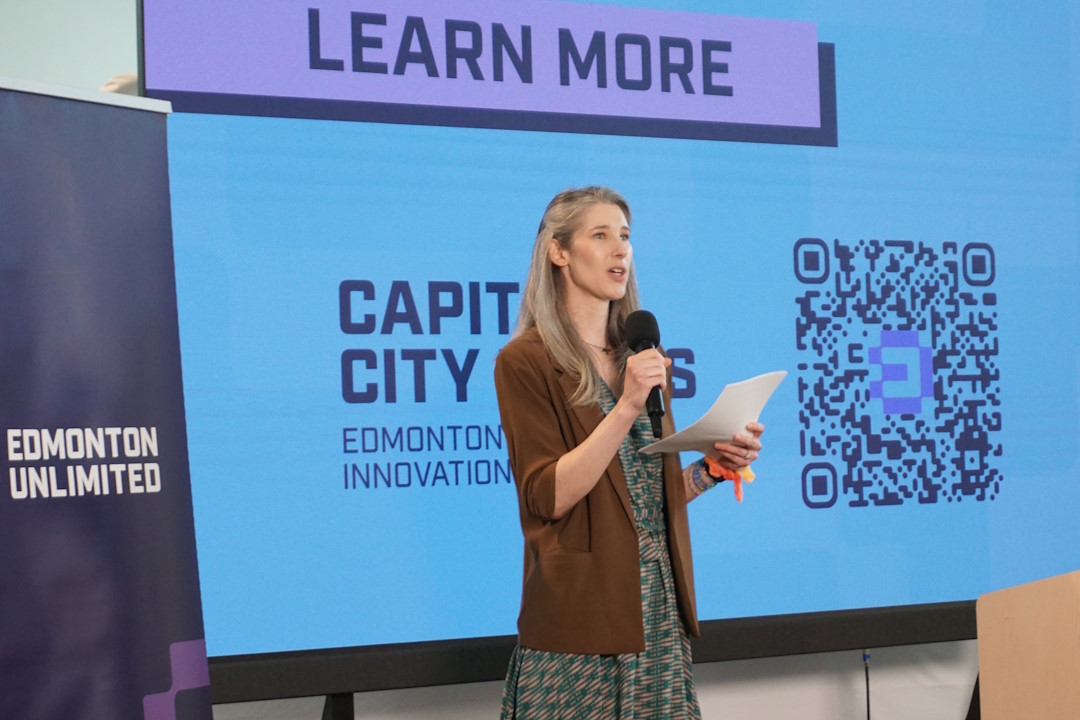 Abbie Stein-MacLean stands in front of a projection screen that reads "Learn More" and "Capital City Pilots" beside an "Edmonton Unlimited" banner