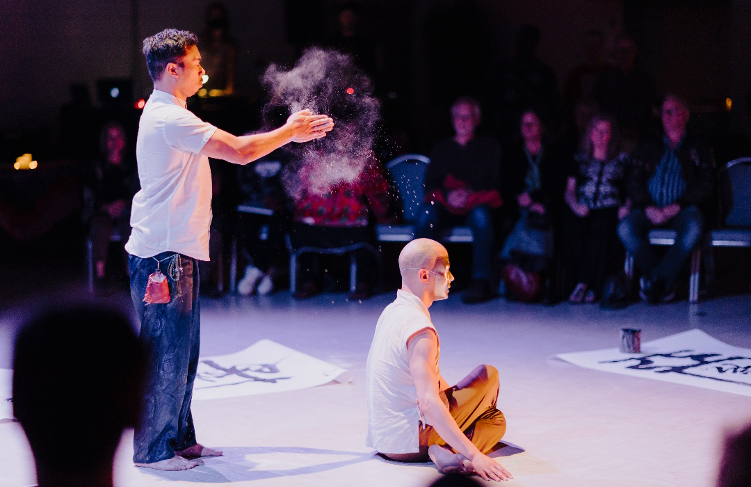 A man claps his hands creating a cloud of dust behind a masked man seated cross-legged on the floor.