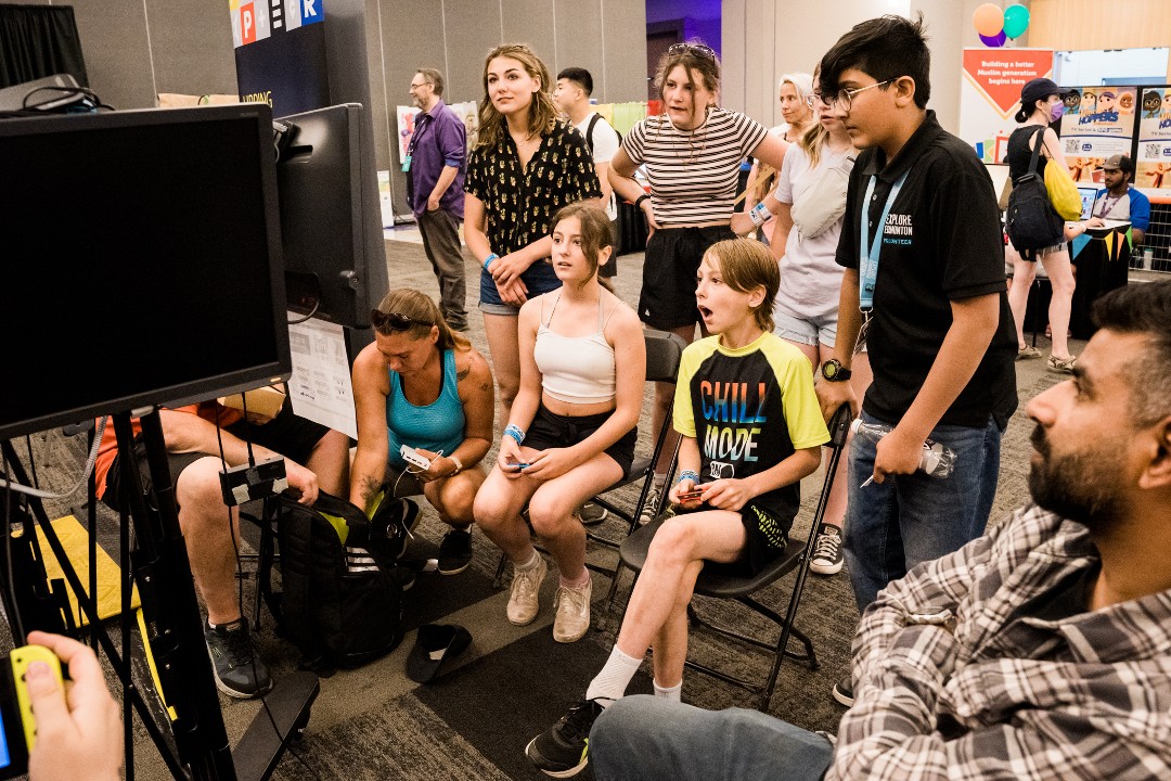 Two young people hold video-game controllers and react to what's on the monitor while others and watch inside an exhibition hall.