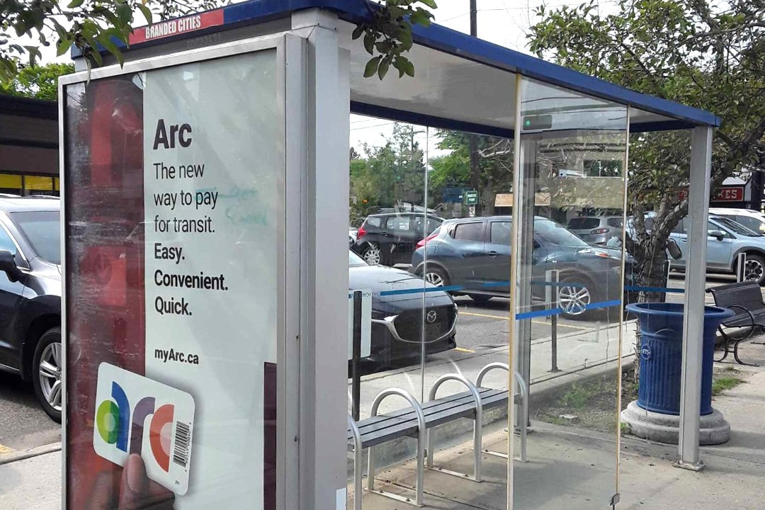 A bus shelter on 124 Street with some light graffiti on an advertisement for the city's Arc card, in front of a parking lot and an overcast sky.