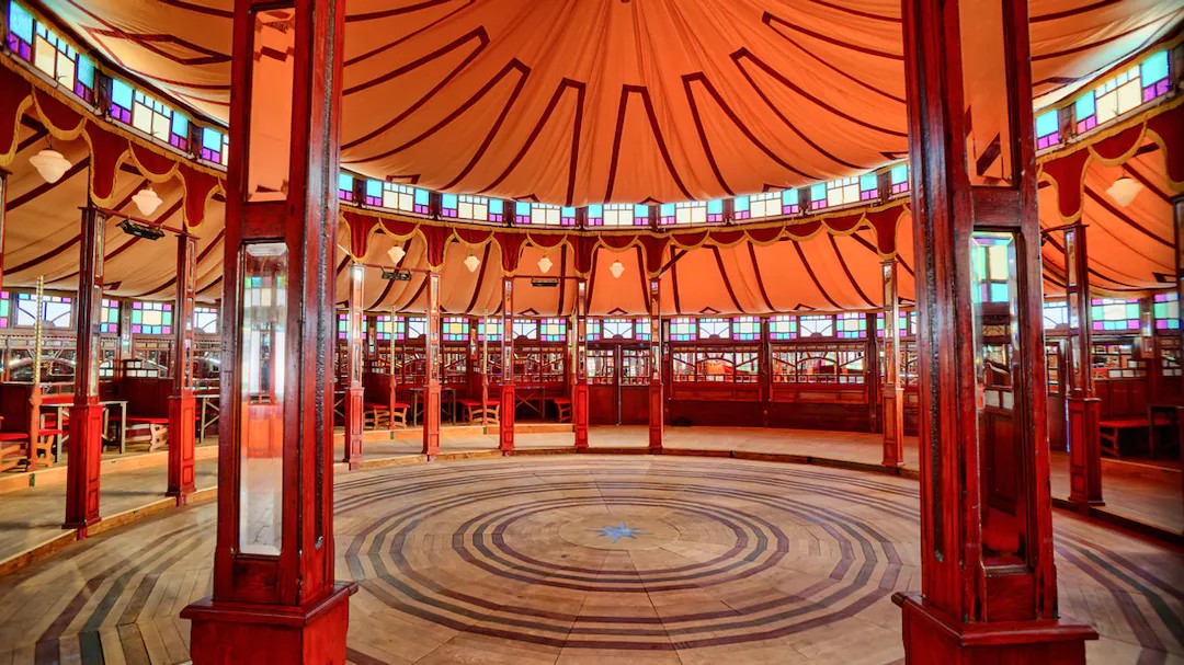 The inside of a bigtop-style tent, with stained glass and mirrors around the edges and a circular stage in the centre