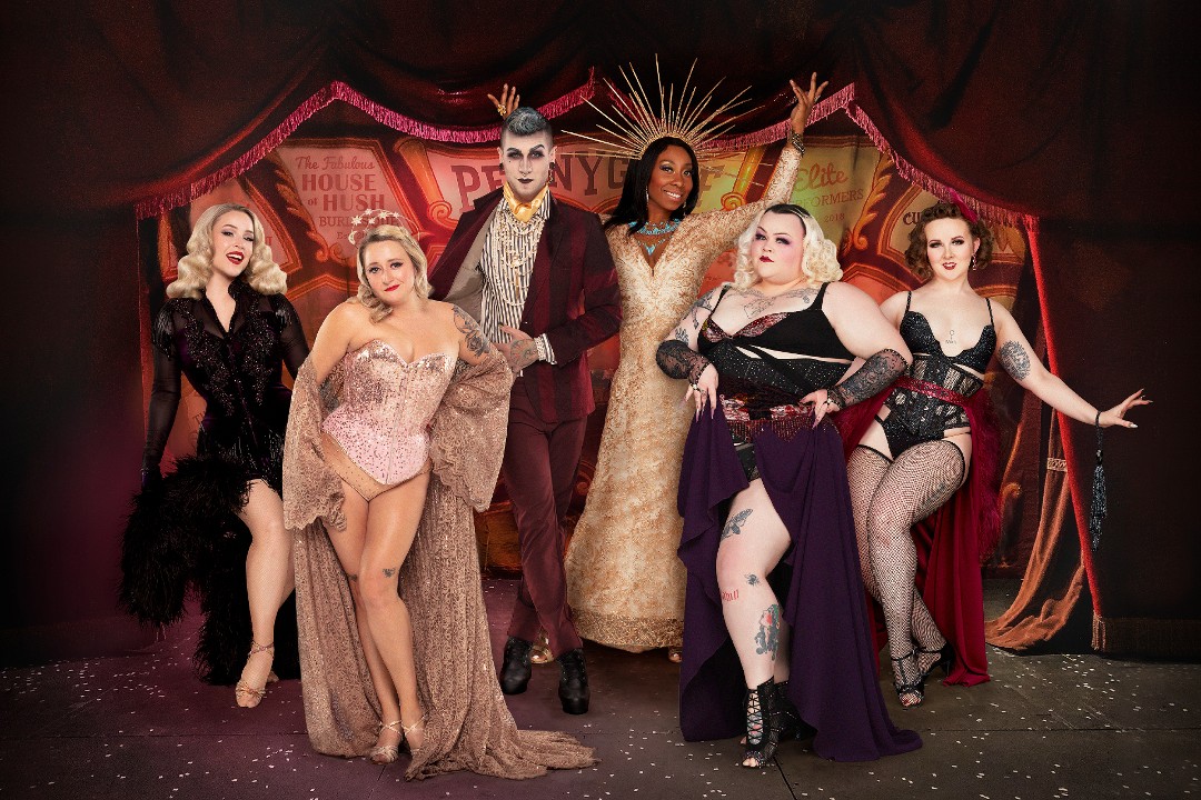 Six burlesque performers pose in costume in front of the opening to a tent and carnival signs.