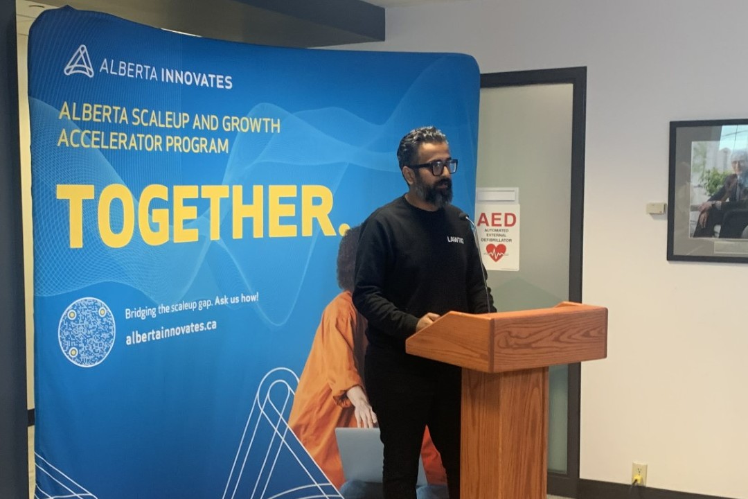 Ali Salman stands at a wooden podium in front of an Alberta Innovates backdrop that reads "Alberta Scaleup and Growth Accelerator Program: Together."