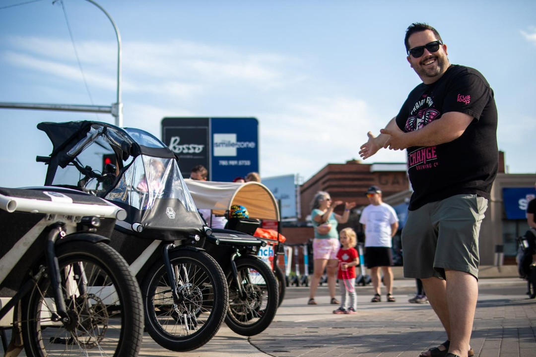 Coun. Michael Janz, dressed in sandals, shorts and a T-shirt, gestures at a row of four electric cargo bikes.