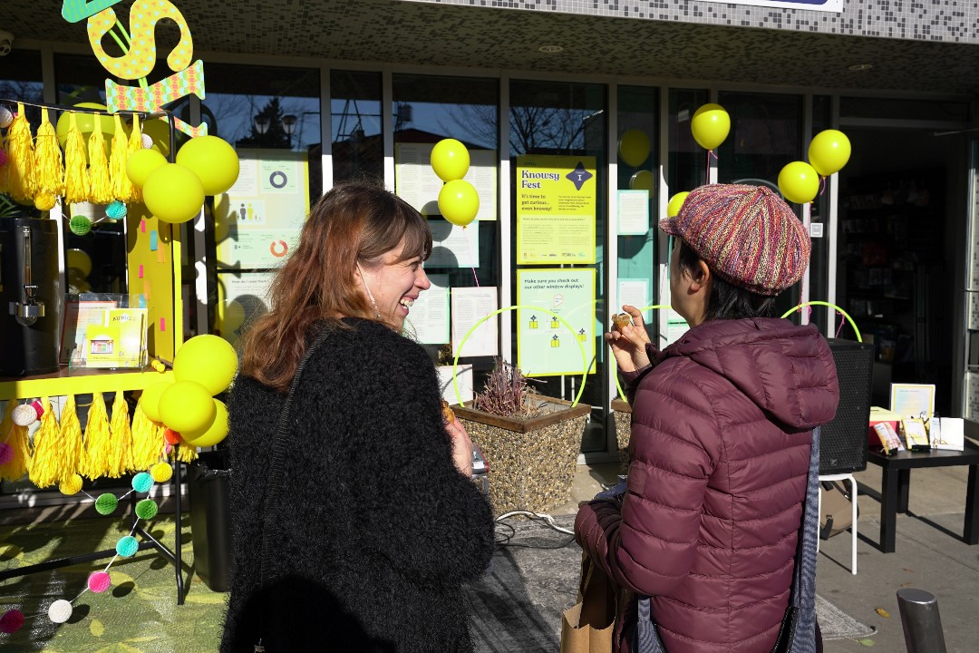 A woman smiles at another person outside of a storefront decorated with balloons, with a sign for Knowsy Fest on the door