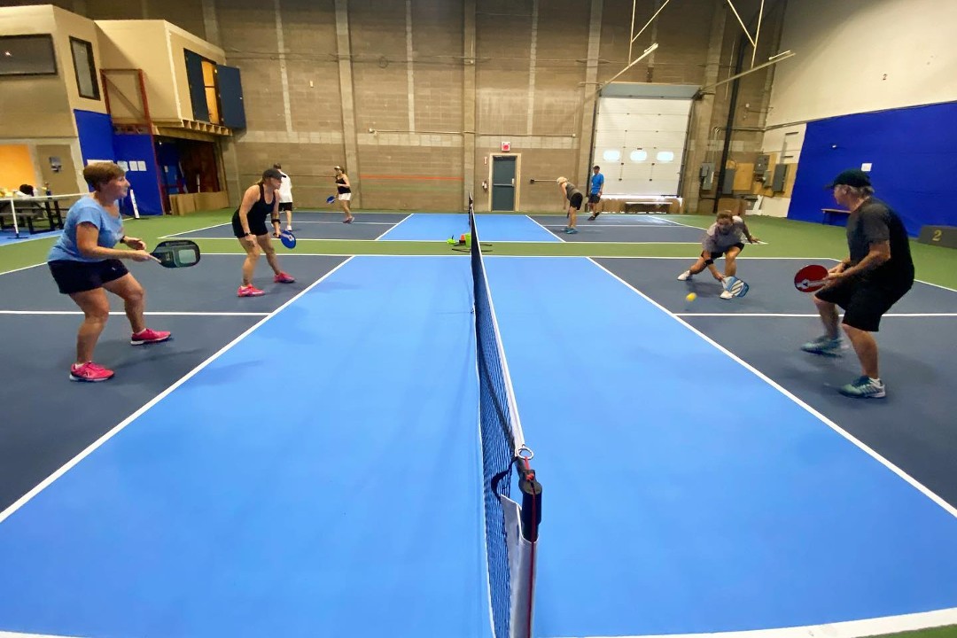 Players face-off on an indoor pickleball court coloured blue, green, and brown. The court's net runs through the centre of the photo, with players holding paddles on either side.