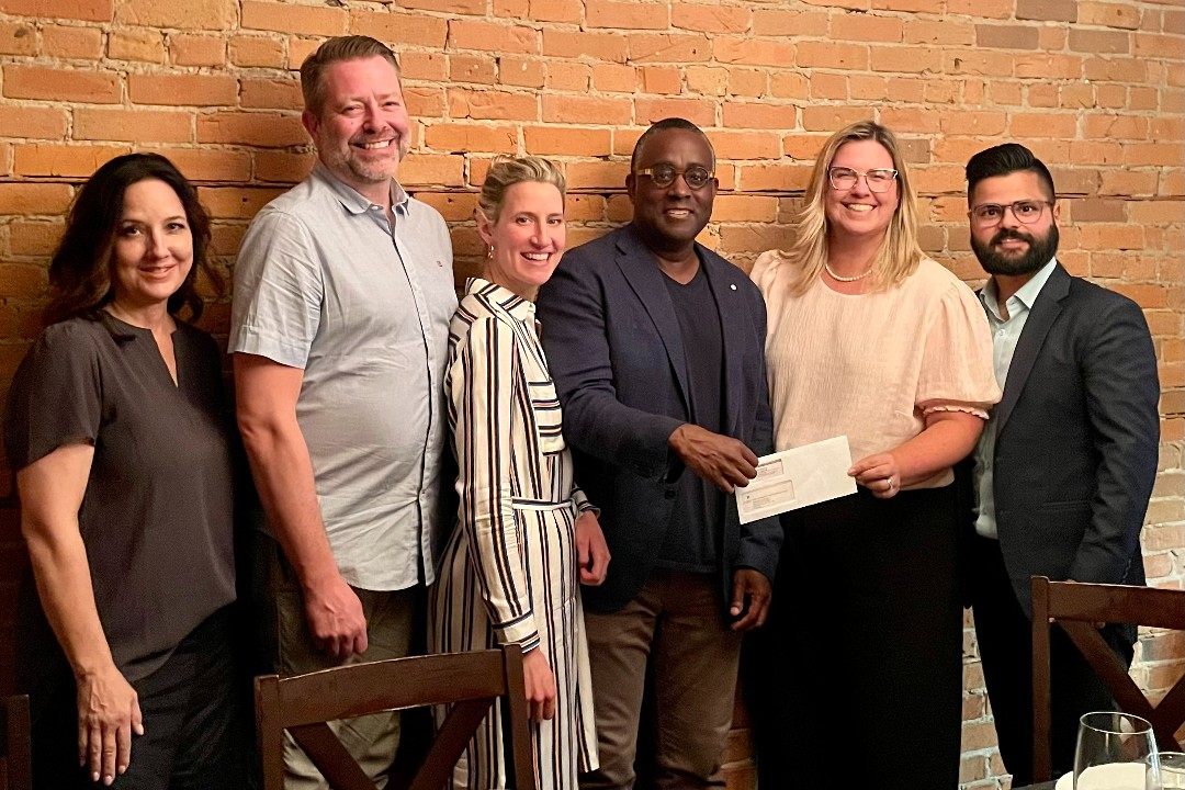 Six smiling people stand against a brick wall; among them are Brennan Walters of Theragraph and Jacqueline Sartor of Pfizer holding an envelope with a cheque in it