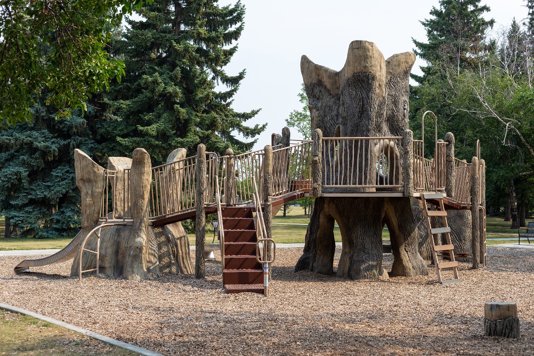 A play structure made of faux logs with stairs, ladders, and a slide, surrounded by pea gravel