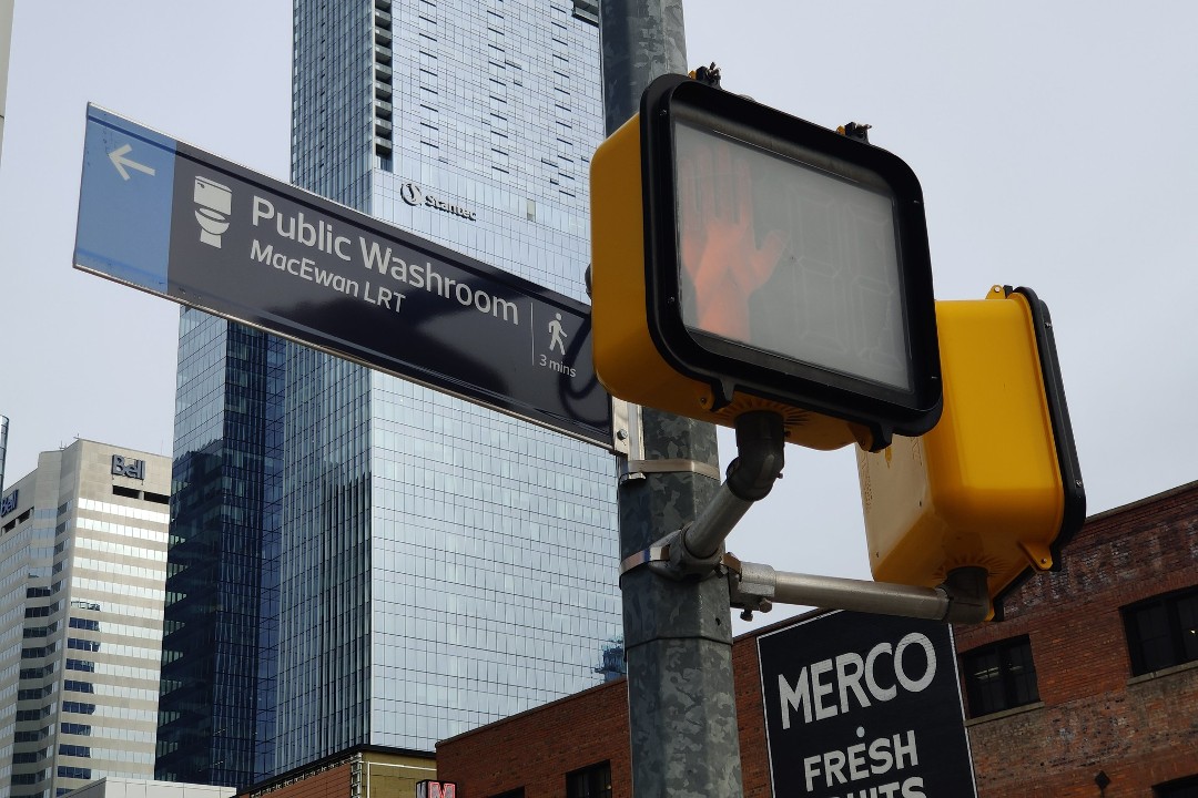 A wayfinding sign pointing to the MacEwan LRT station washroom, fixed to a downtown street pole next to a pedestrian signal displaying an orange hand