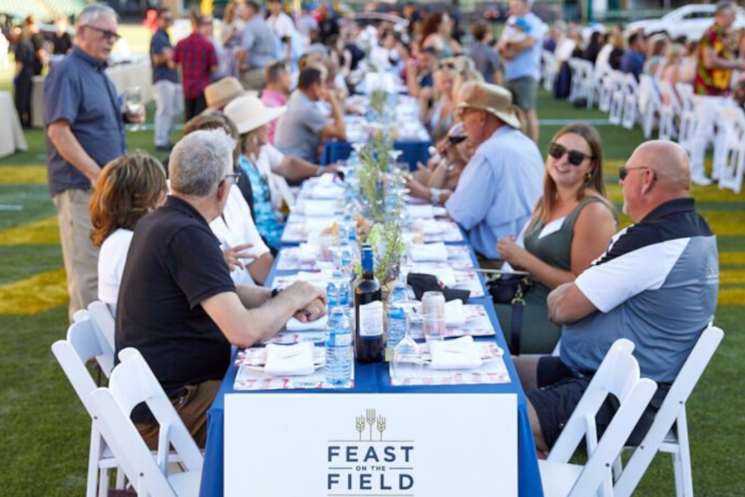 People sit at several long dining tables outdoors, with a sign reading "Feast on the Field"