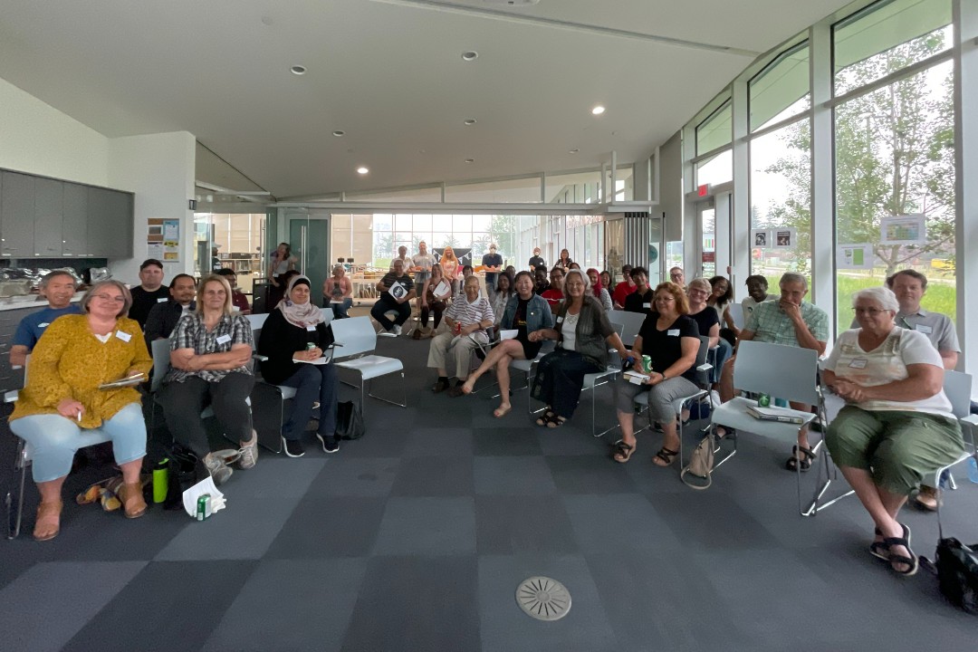 A large group of people is seated on chairs inside a white-and-grey space with large windows.