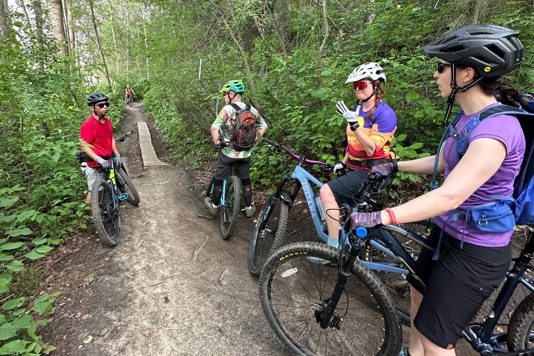 Four mountain bikers stop on a trail while another approaches on the other side of a boardwalk on a trail in a wooded area