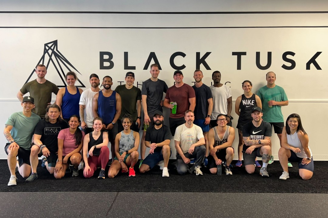 Twenty-one smiling people in exercise gear stand in two rows in front of a wall with the sign Black Tusk