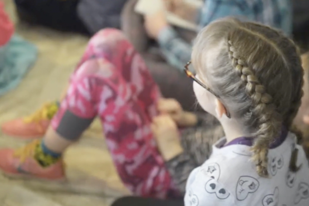 A child with blonde braids and tortoise glasses sit with other children the floor.