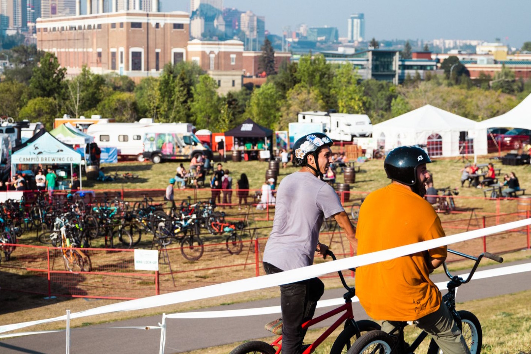 Two people on mountain bikes look down at a field of tents and bikes, with the Rossdale plant in the background