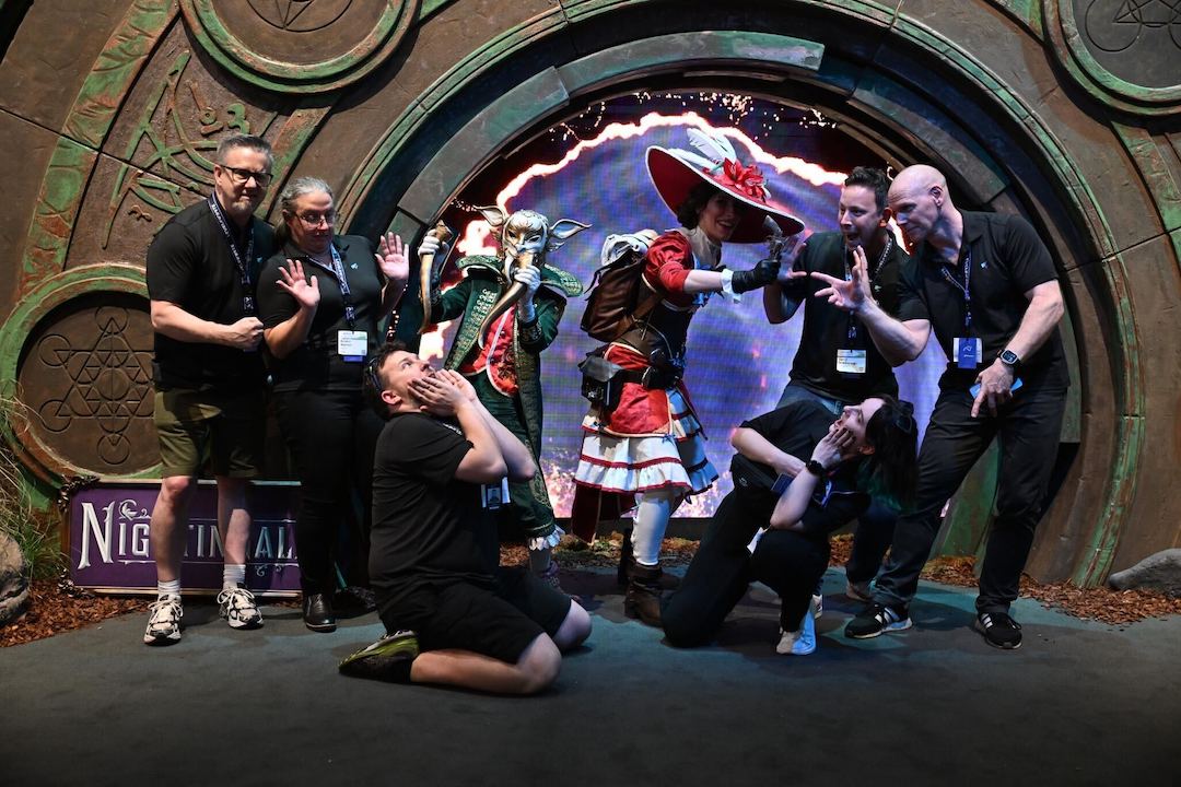 Two cosplayers — a fae in a long coat with epaulettes and a lady in a knee-length frock and a big, fancy hat — pose with six black-clad conference-goers in front of a portal promoting Nightingale.