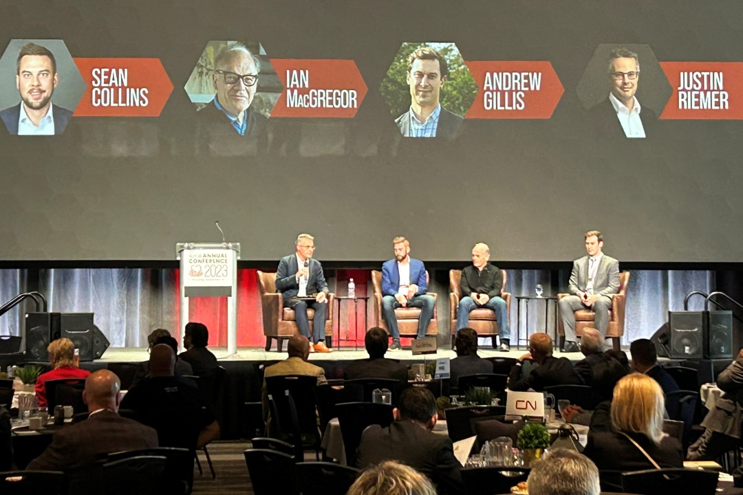 Justin Riemer, Sean Collins, Ian MacGregor, and Andrew Gillis sit on a conference stage beneath a screen showing their names