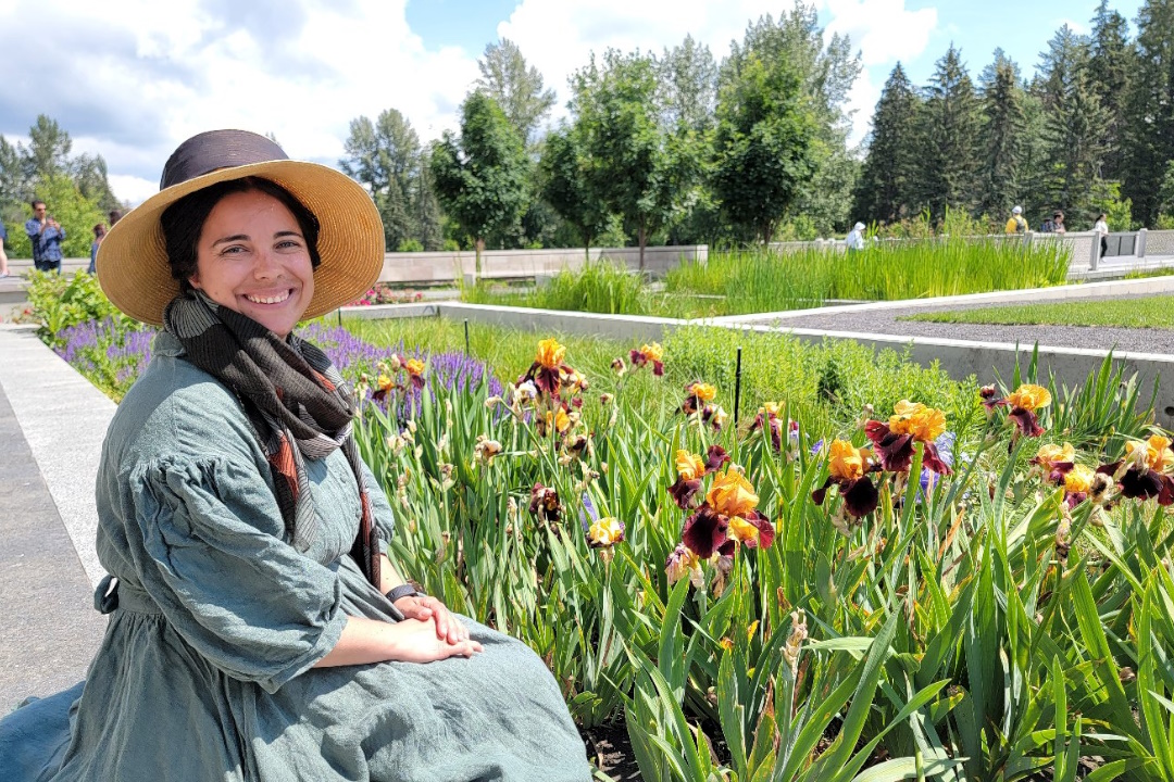 A smiling woman in a floppy hat and denim dress sits beside some flowers