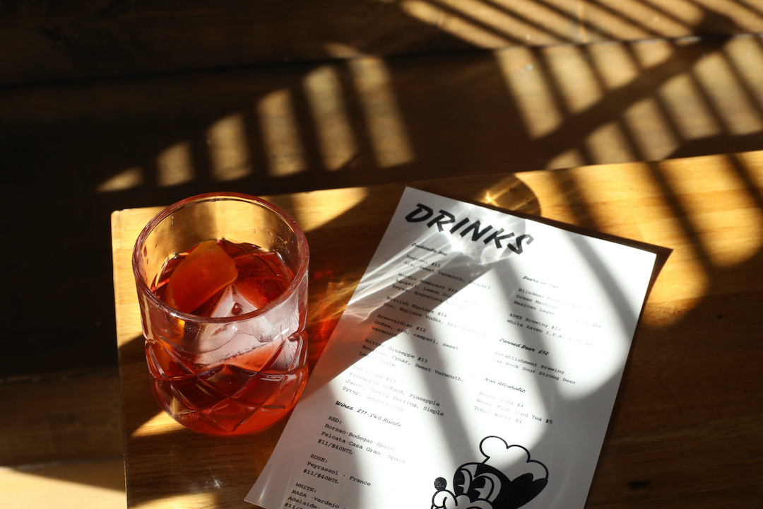 A red cocktail sits in a sunbeam on a wooden table beside a menu with the heading "DRINKS".