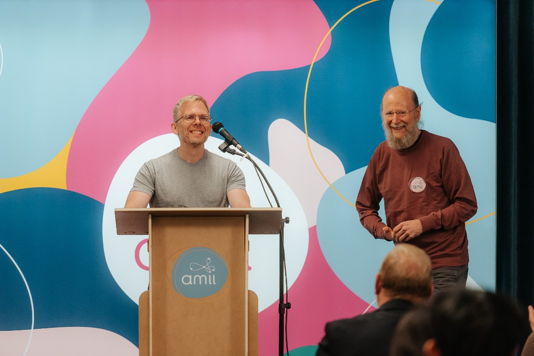 A smiling John Carmack stands behind a podium with the Amii logo on the front, while Rich Sutton, wearing an Amii sweater, smiles beside him