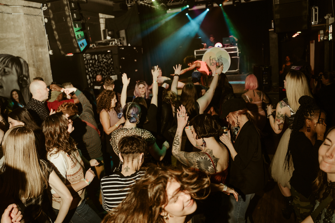 Around 20 people are dancing in front of a stage that has a DJ behind a Fruit Loop logo on it.