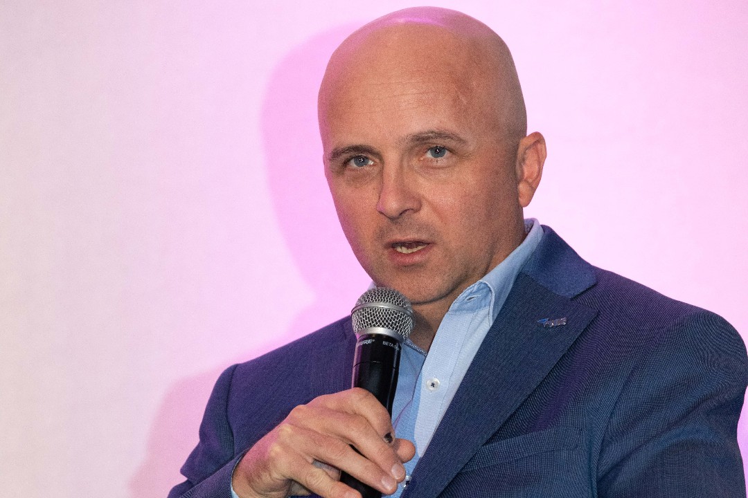 A close-up of a bald person holding a microphone and wearing a blue suit, light in front of a white background by pink-ish light.